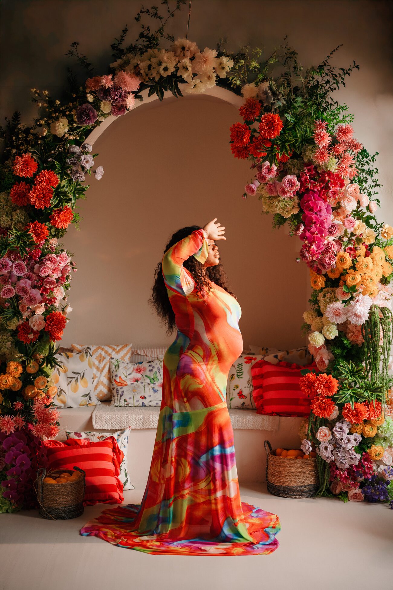 Expectant mother in a flowing rainbow gown beside a floral arch
